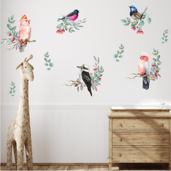 Decorative wall stickers of birds and flowers on a wall with a wooden dresser and stuffed giraffe in the foreground.