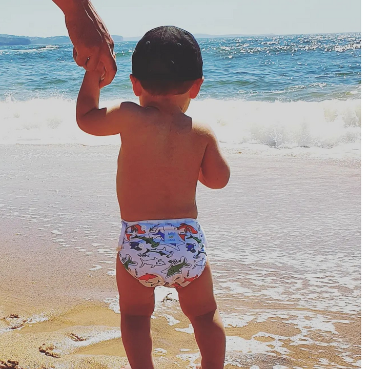 A toddler standing at the beach wearing a swim nappy with a shark design, holding hands with an adult.